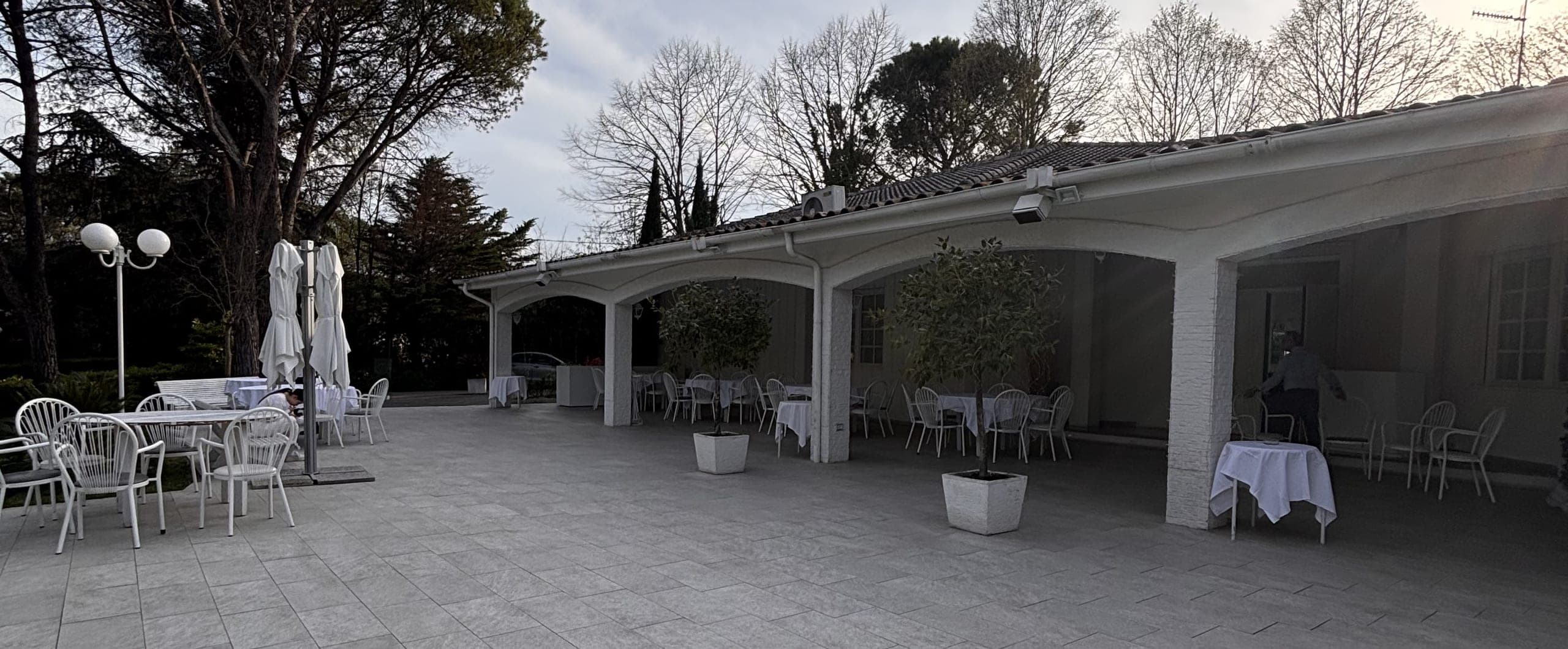 La terrazza della villa con i tavolini bianchi, il portico ad archi e i pini del parco